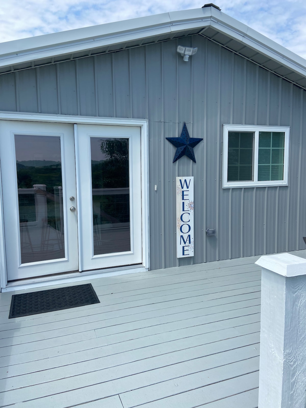 The entrance of the apartment is presented, showcasing a spacious deck with light-colored flooring. Double glass doors lead inside, accompanied by a decorative blue star and a welcoming sign. A small window allows natural light into the entryway.