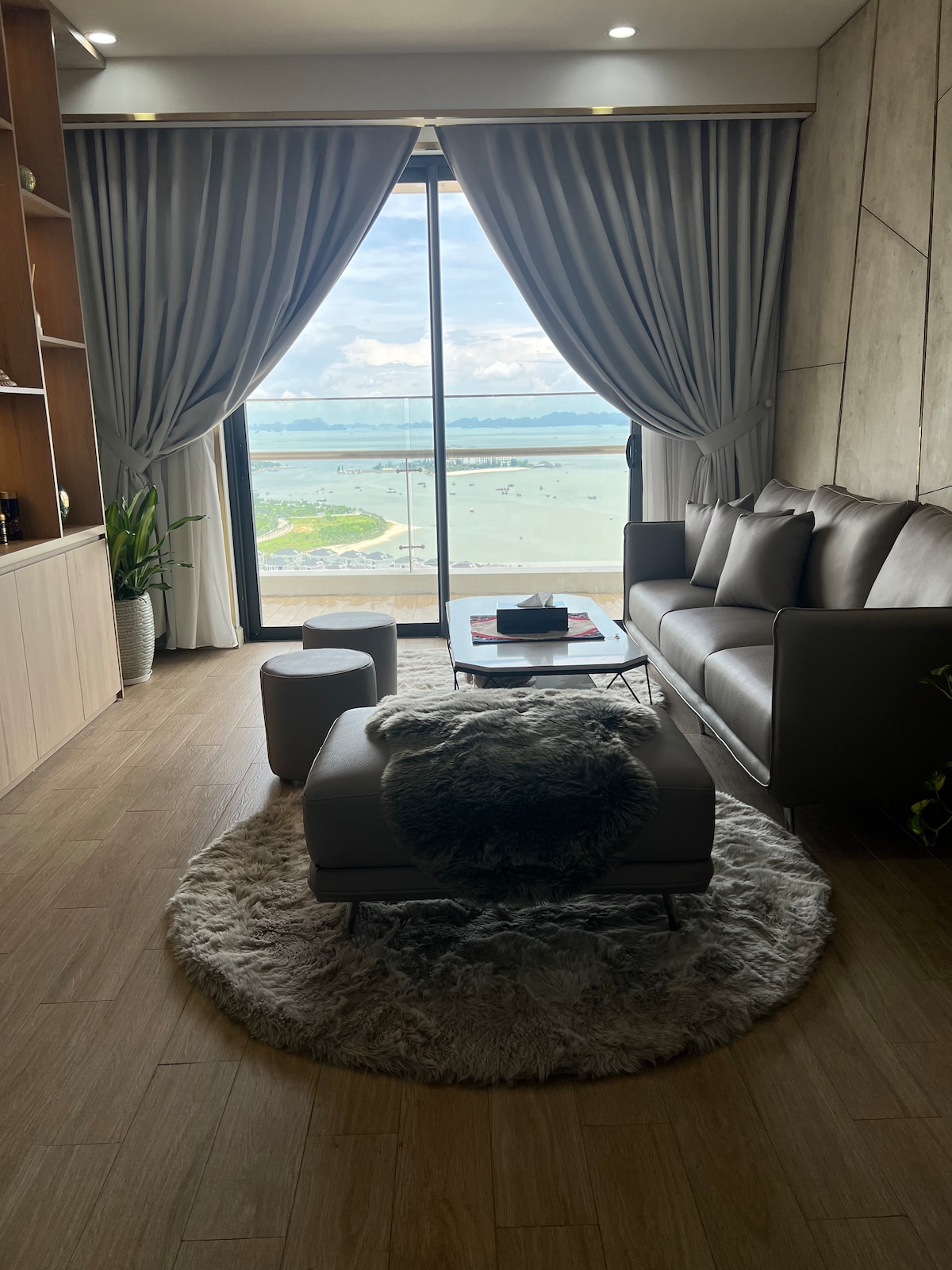 A living room is presented with large windows overlooking a serene sea view. A comfortable sofa is positioned near a round coffee table on a soft area rug. Decorative plants add a touch of greenery beside the floor-to-ceiling curtains.