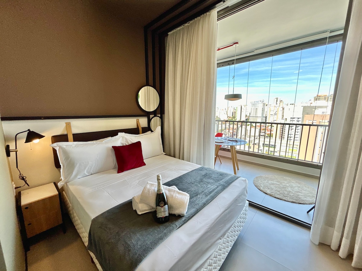 A well-appointed bedroom features a spacious bed adorned with white linens and a dark throw blanket. A small round table with two chairs stands near the large glass window, revealing a cityscape view. Soft curtains frame the window, enhancing the room's natural light.