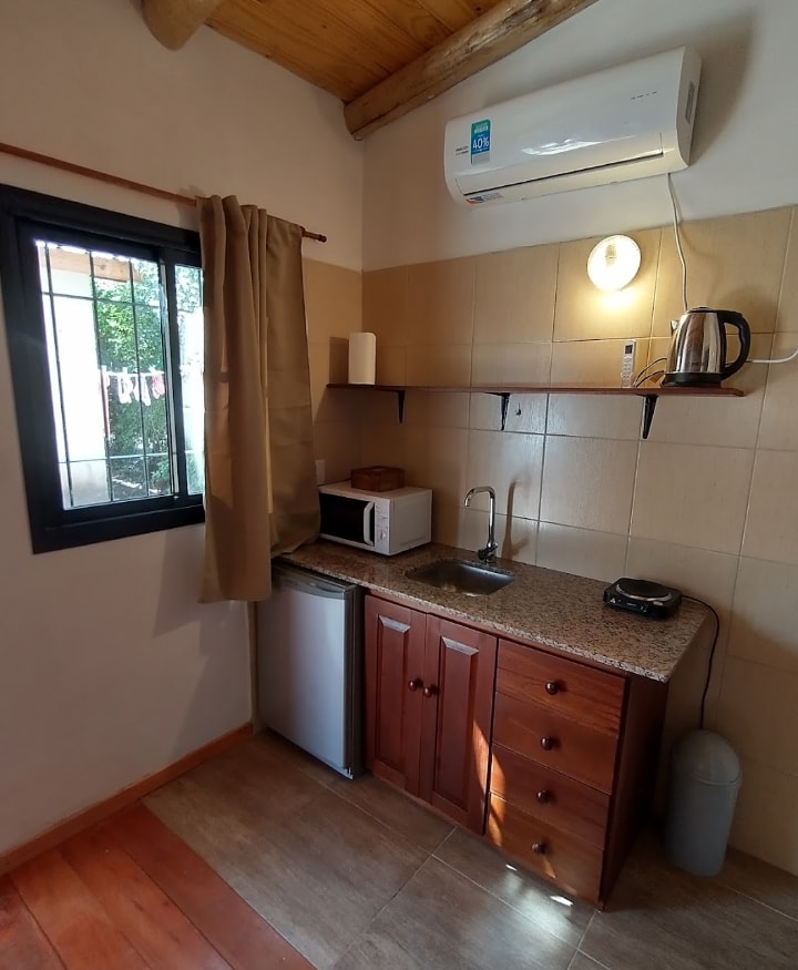 A compact kitchen area is equipped with essential appliances, including a microwave, kettle, and a small fridge. Wooden cabinetry provides storage space, while a stone countertop offers a work area. Natural light enters through the window, complemented by soft curtains.