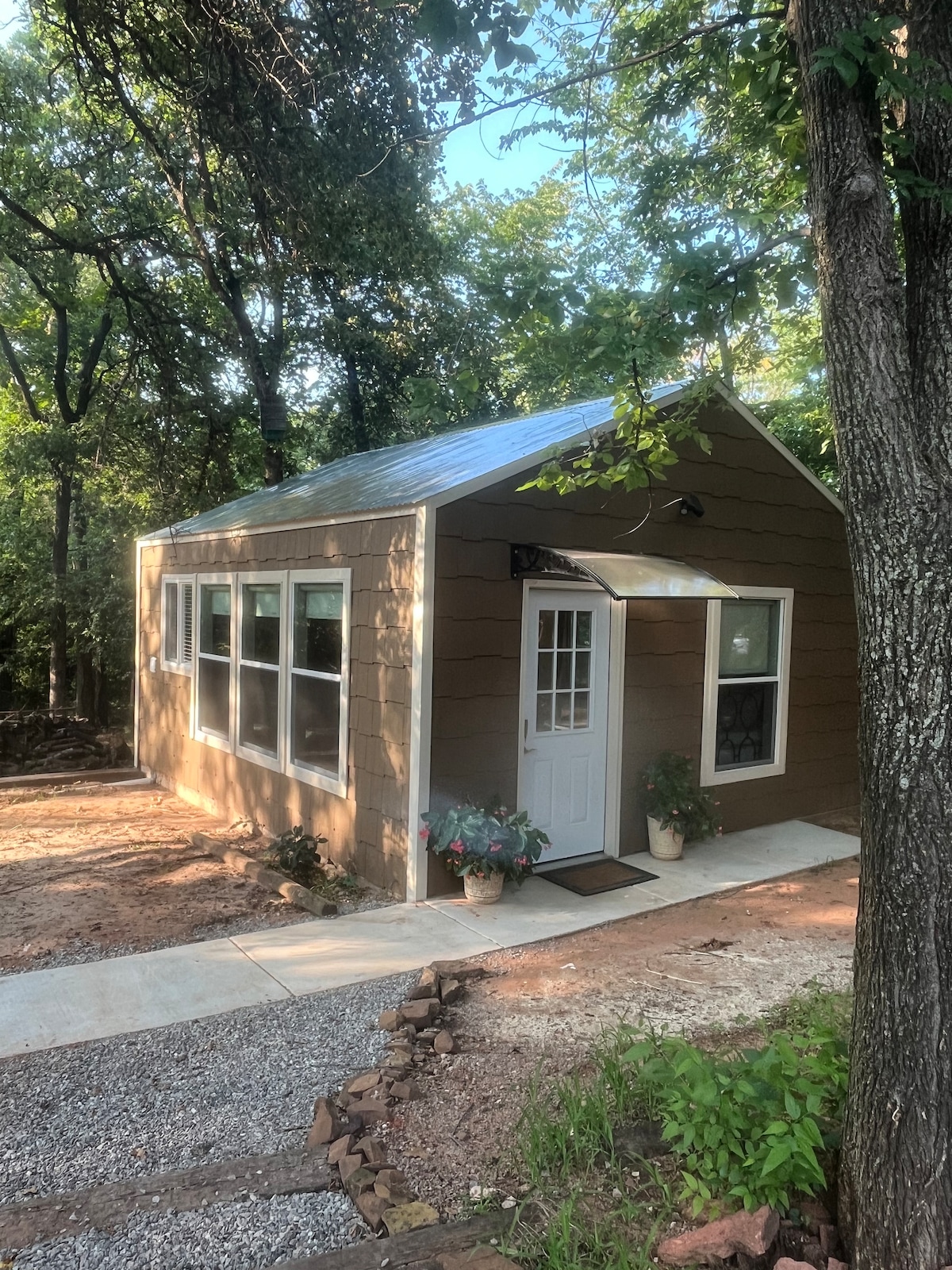 A charming cottage exterior is depicted, featuring a sloped roof and large windows that allow natural light to enter. The entrance includes a front door framed by flowers in pots, with a pathway lined with gravel leading to the door amidst a surrounding wooded area.