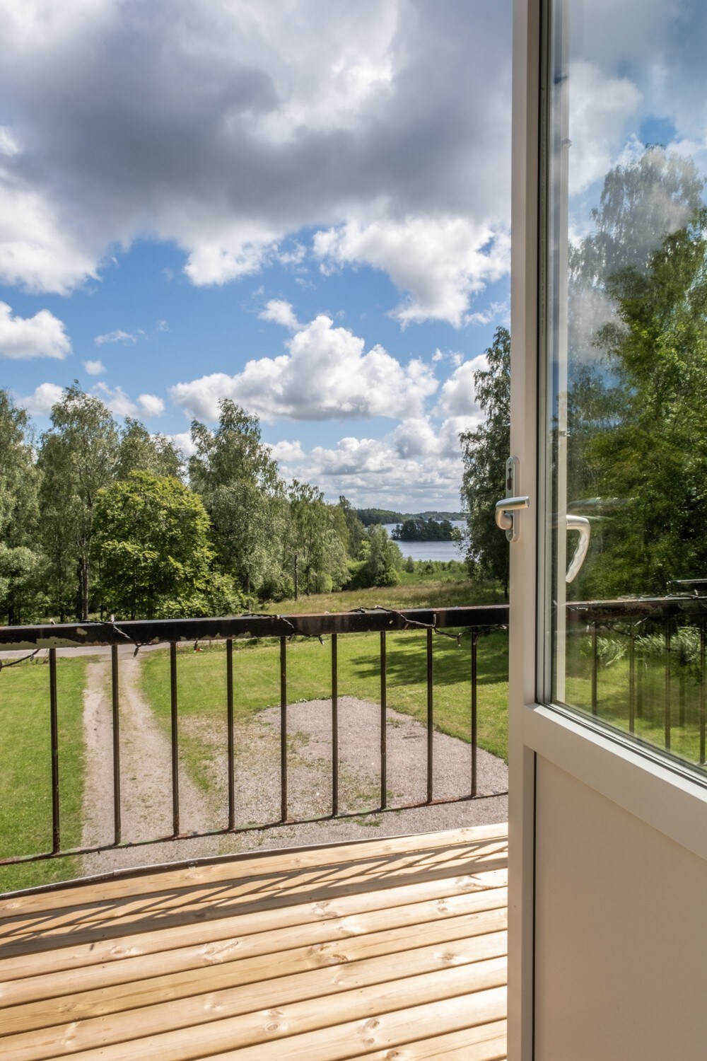 A view from the balcony is presented, featuring a pathway that leads through a green landscape towards a visible body of water. The sky is partly cloudy with sunlight illuminating the scene. The railing and wooden floor of the balcony frame the picturesque outdoor setting.