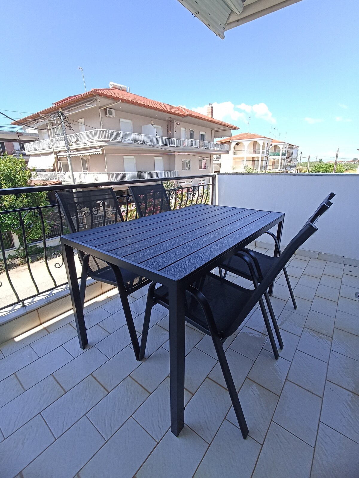 A balcony area features a black metal table surrounded by two matching chairs. The space is bathed in natural light, with a view of nearby buildings and greenery visible beyond the railing. The tiled flooring adds a touch of elegance to the outdoor setting.