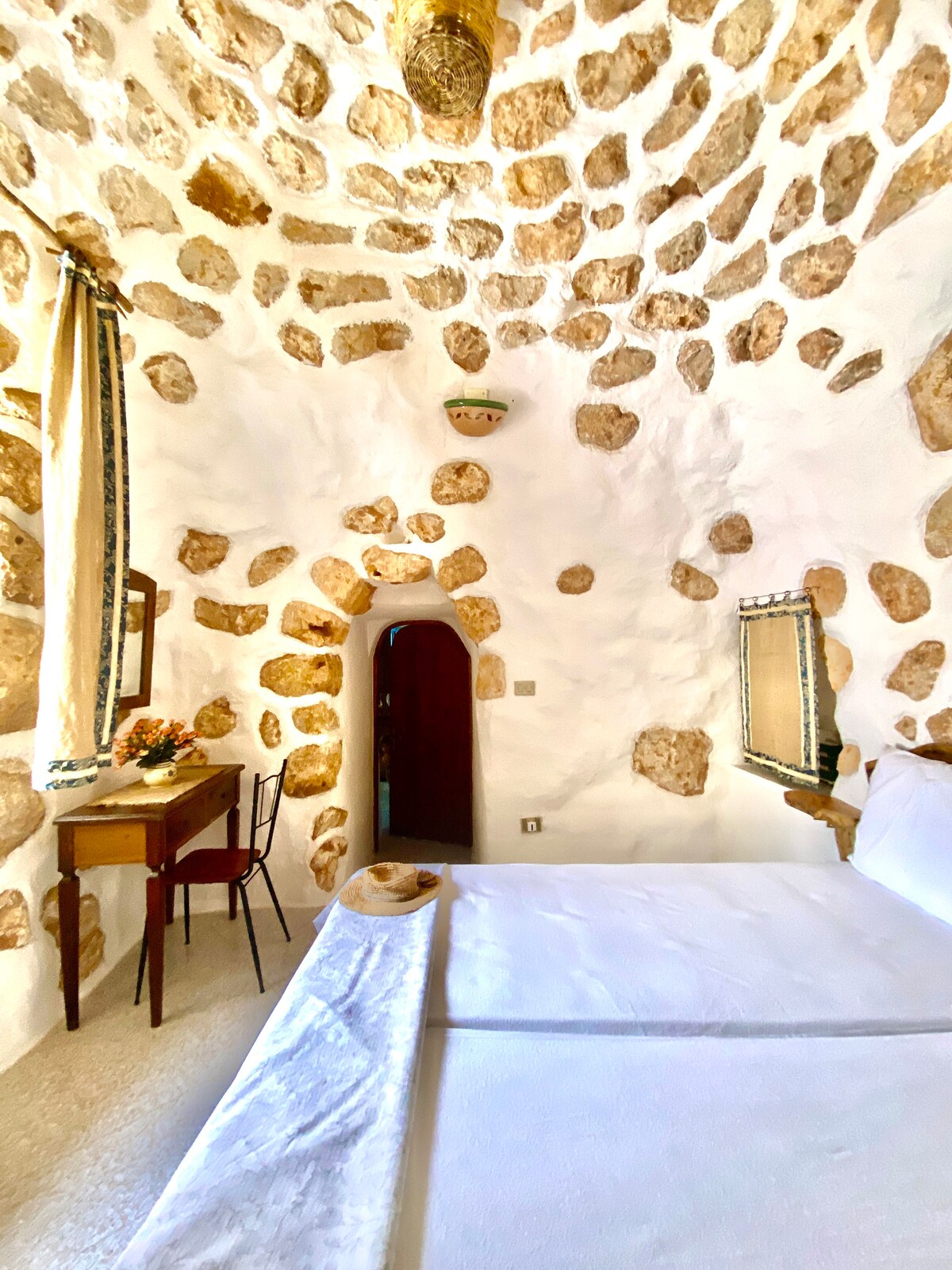 A traditional trullo features rounded stone walls and a high domed ceiling, creating a unique sleeping area. A spacious bed is positioned alongside a small wooden table and chair. A simple mirror and a decorative light fixture add to the room's charm.