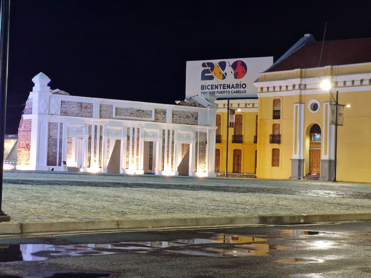 An illuminated building facade is showcased at night, featuring exposed brick and architectural details. A large sign reading 'BICENTENARIO 200' is prominently displayed, with reflections visible in nearby wet pavement. The neighboring structure complements the scene with its historical yellow exterior.