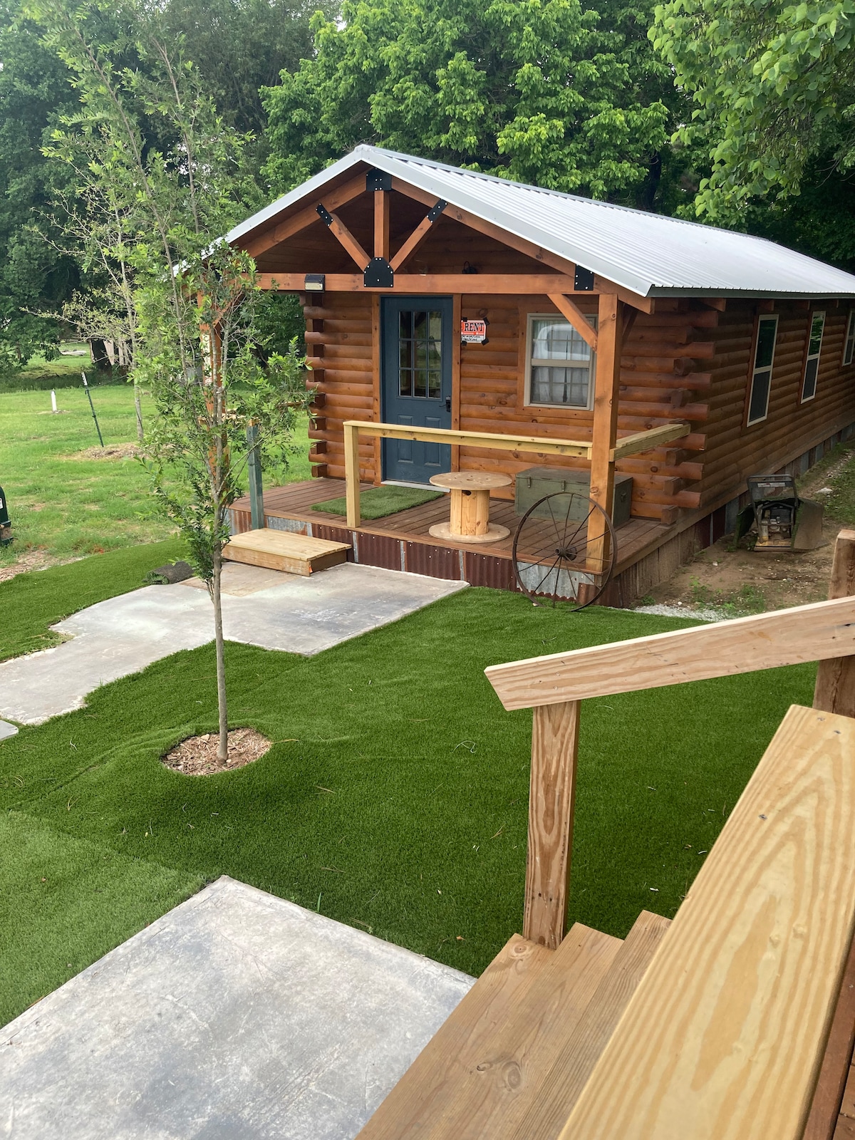 A charming log cabin is nestled amidst greenery, featuring a covered porch and a round wooden table. A stone pathway leads to the entrance, while freshly manicured grass surrounds the cabin, inviting outdoor relaxation.
