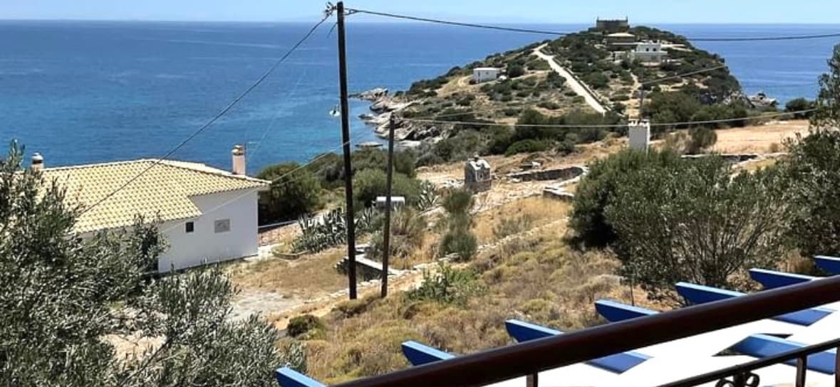 A view from the balcony showcases rolling hills leading to the coastline, where turquoise waters meet the rocky shore. A single-story house is positioned in the foreground, with olive trees scattered throughout the landscape. Power lines add a practical touch to the serene setting.