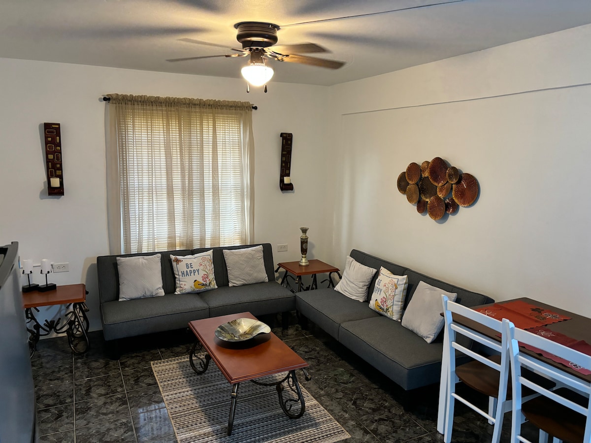 A spacious living area features two gray sofas adorned with decorative pillows and a coffee table. A light rug enhances the tiled floor, while a ceiling fan provides airflow. Natural light filters through the sheer-curtained window, contributing to a bright and relaxed ambiance.
