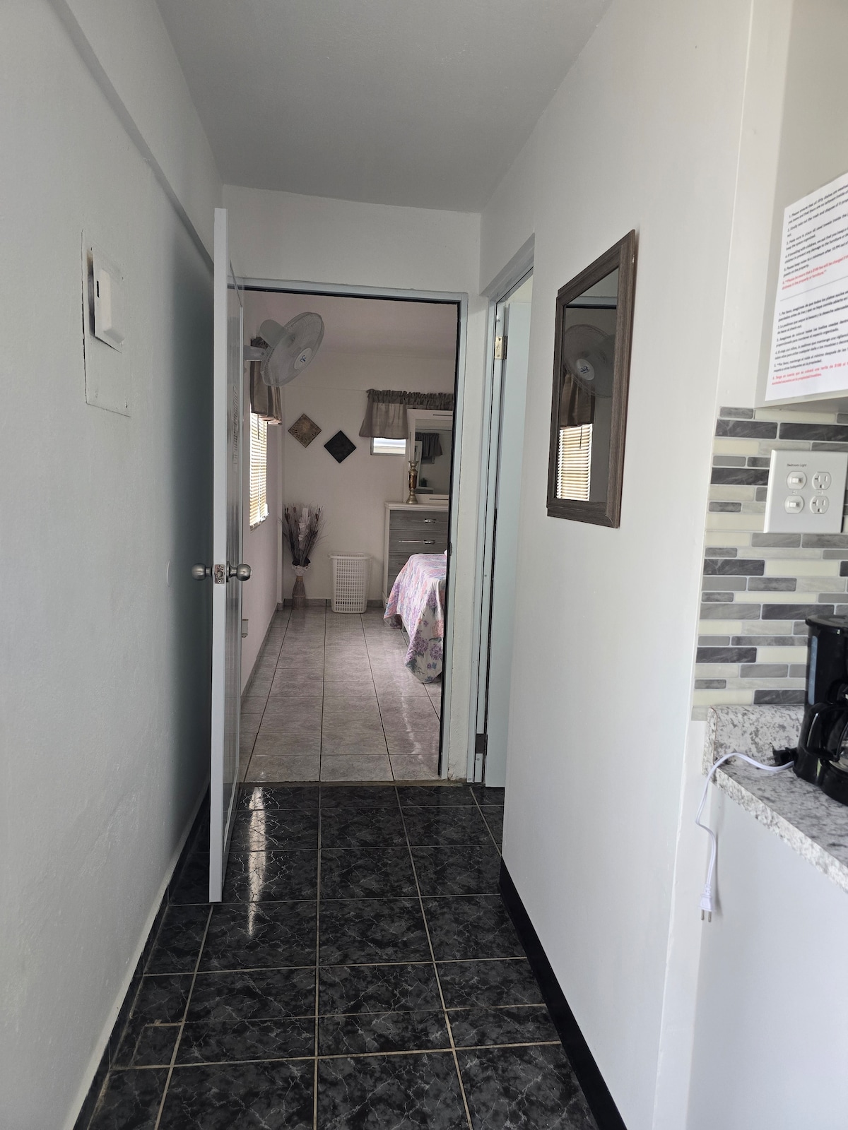 A well-lit hallway leads towards an open bedroom area, featuring light-colored walls and tiled flooring. A mirror hangs on the wall next to the entrance, while a kitchen area with granite countertops is partially visible on the left, enhancing the overall spaciousness.
