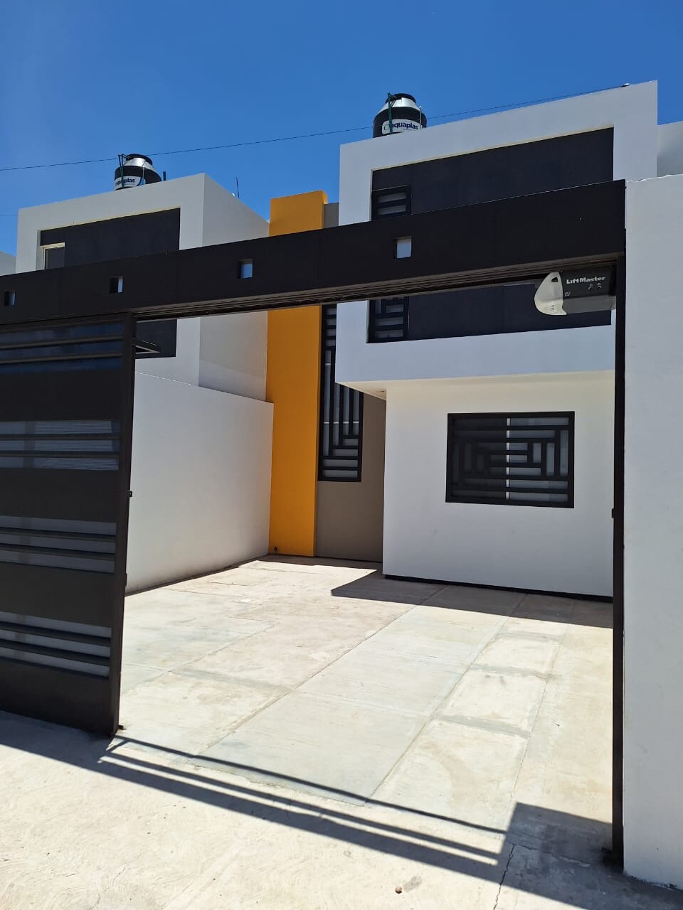 A modern entrance gate opens to a spacious outdoor area, revealing the contemporary facade of the house. Large windows with geometric designs are visible on both sides, complemented by a striking yellow accent wall. The bright blue sky enhances the overall setting.
