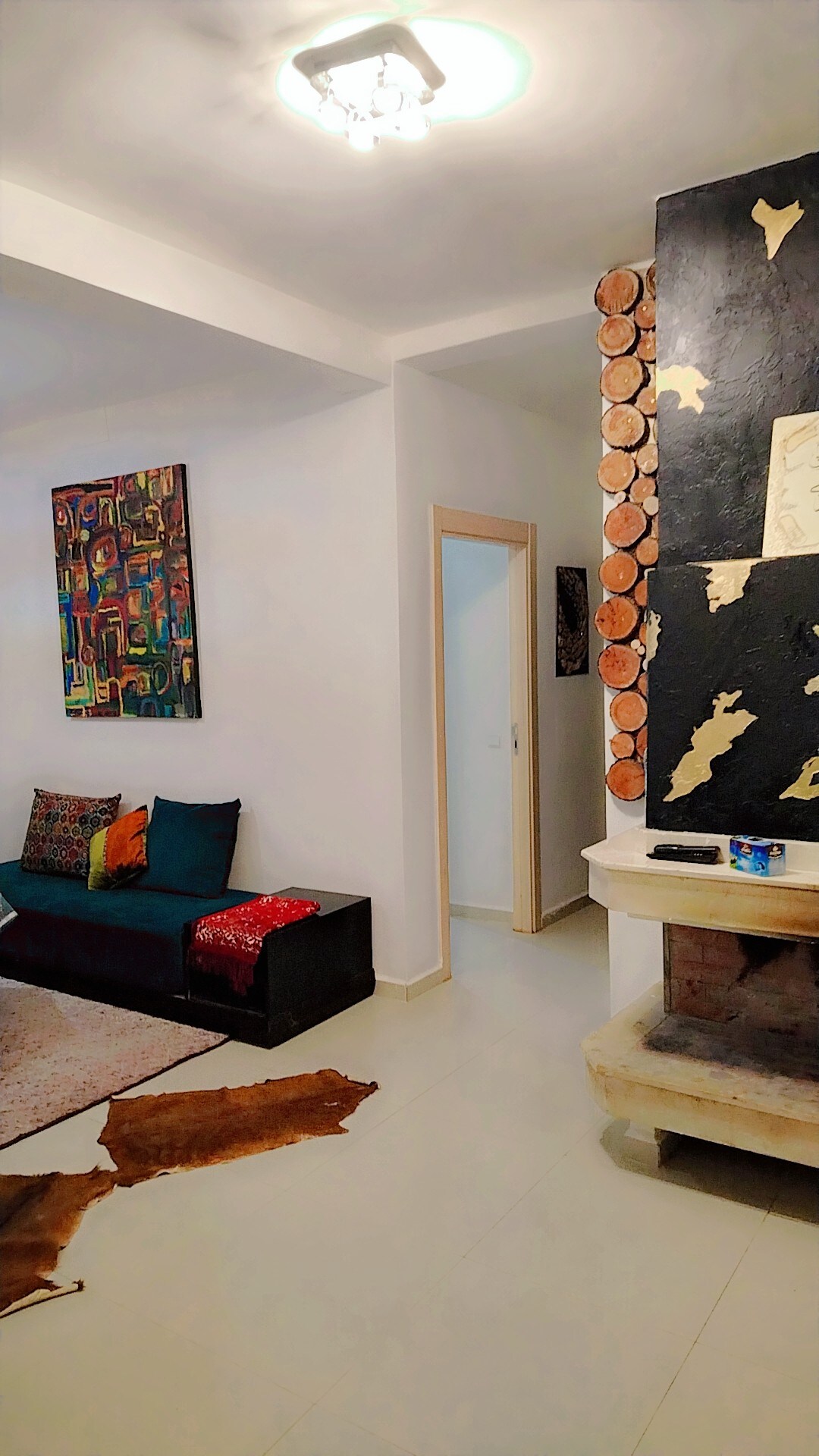 A welcoming living area is displayed with a dark sofa adorned with colorful cushions. A textured area rug is positioned on the floor, partially covered by natural, earthy materials. A modern fireplace is visible along one wall, surrounded by decorative wooden elements and art pieces.