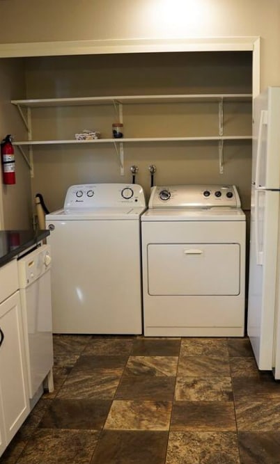 A laundry area contains a combination washer and dryer, with shelving positioned above for storage. The floor features tiles in a natural stone pattern, while appliances are situated near a white refrigerator, contributing to a functional and organized space.