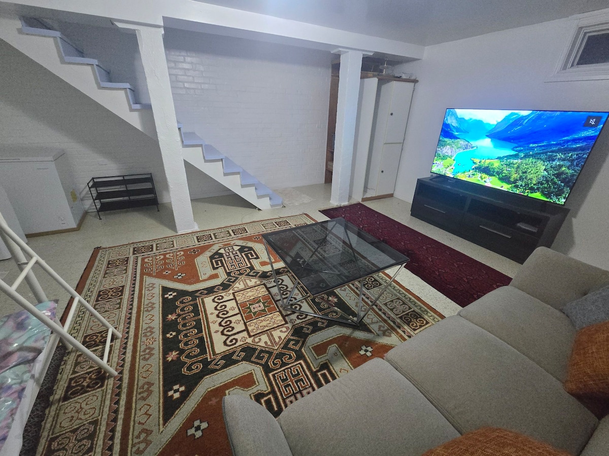 A spacious living area is highlighted by a large television mounted on a dark cabinet. A glass coffee table rests on a decorative area rug, providing a focal point. Soft seating is arranged comfortably, while white stairs lead to the upper level.