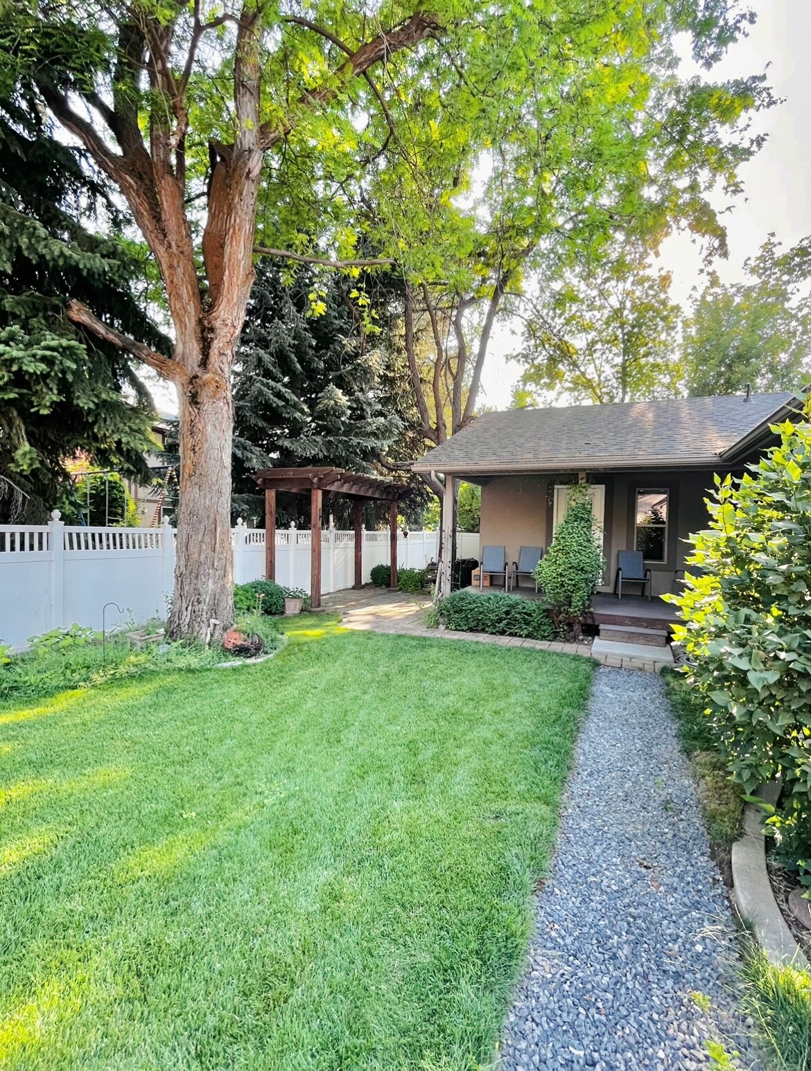A lush green lawn is framed by tall trees, providing a serene atmosphere. Pathways of natural stone and gravel guide towards a covered seating area, while a white fence encloses the yard, enhancing privacy in the inviting outdoor space.