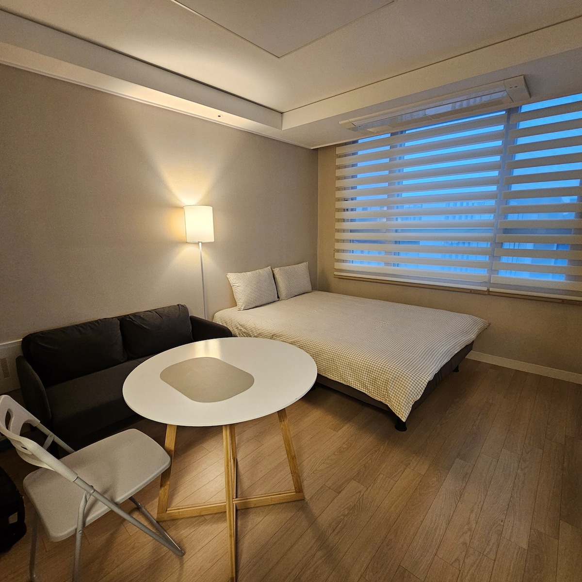 A cozy room features a spacious bed with neutral bedding and two pillows. A sofa is positioned next to a small round table with golden legs. The window is covered with horizontal blinds, allowing soft light to enter the space.