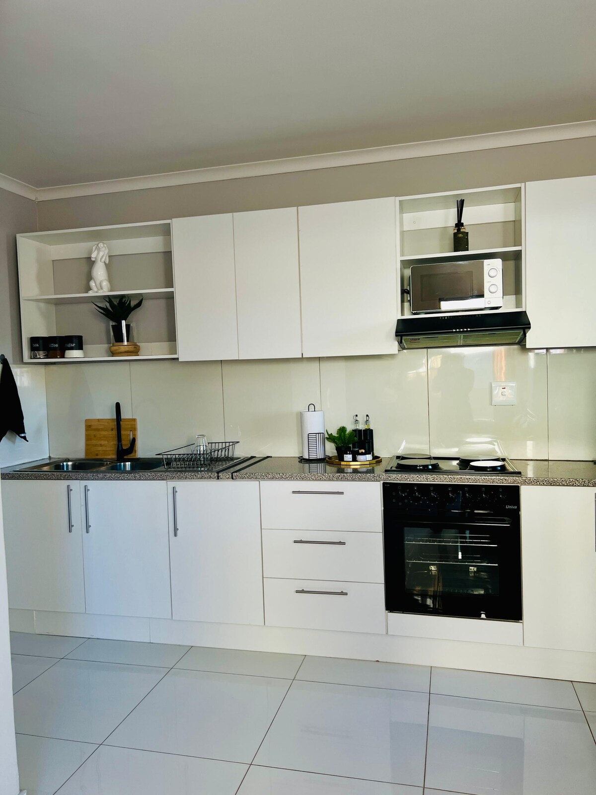 A modern kitchen features sleek white cabinetry and a glossy countertop. Above, open shelves display decorative items and a small plant. Appliances include a microwave, stove, and oven, while a dish rack and utensils are neatly arranged on the counter.