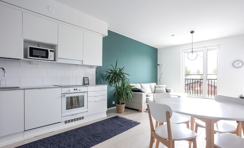 A modern kitchen area includes sleek cabinetry and essential appliances such as a microwave and oven. A dining table with white chairs is positioned near a cozy living area featuring a light-colored sofa and a plant. Large windows allow natural light to fill the space.