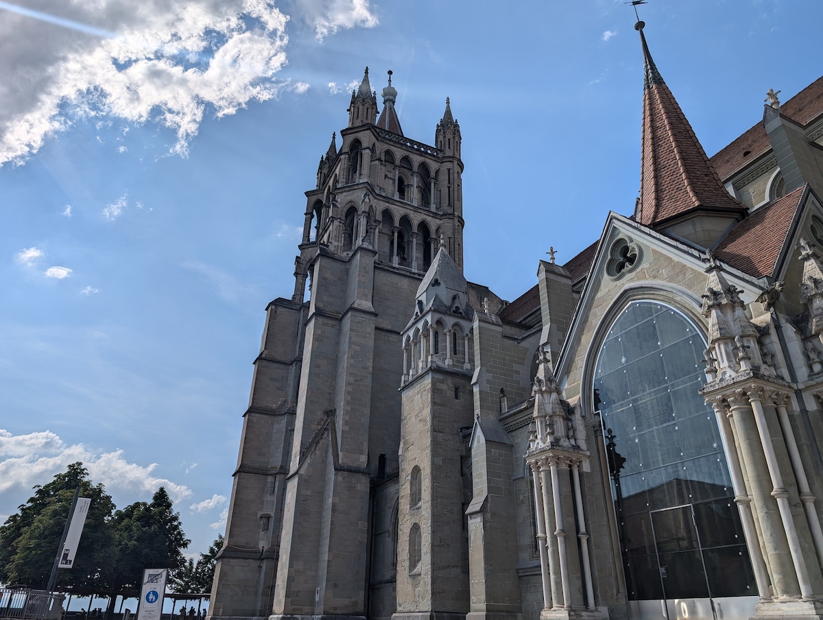 A prominent church building features a mix of architectural styles with tall towers and detailed stonework. Large windows are visible, reflecting the sky, while pointed roofs add elegance to the structure. Green trees can be seen in the foreground, contributing to the serene atmosphere.