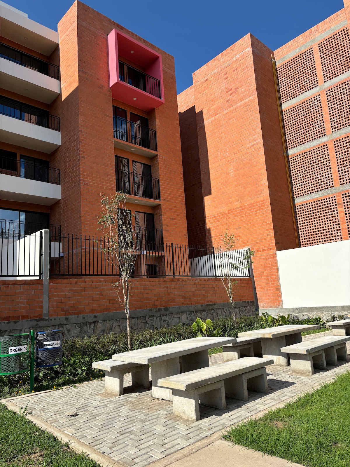 A modern apartment building with a combination of red brick and colorful accents is shown. In the foreground, there are several concrete benches arranged in a landscaped area, surrounded by small plants and greenery. The building features balconies, enhancing its architectural appeal.