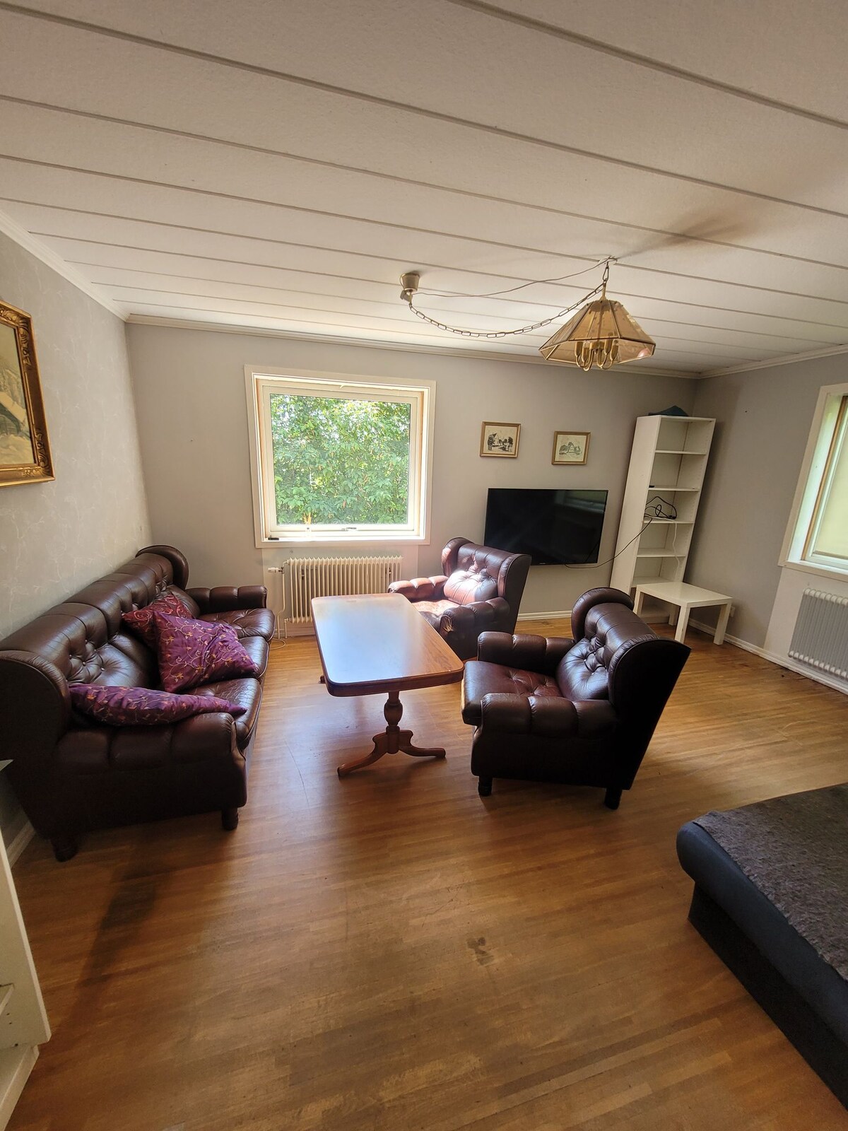 A comfortable living area features two dark leather sofas facing each other, with a wooden coffee table in between. Natural light enters through a window, illuminating the space. A television is mounted on the wall, and a white bookshelf stands in the corner.