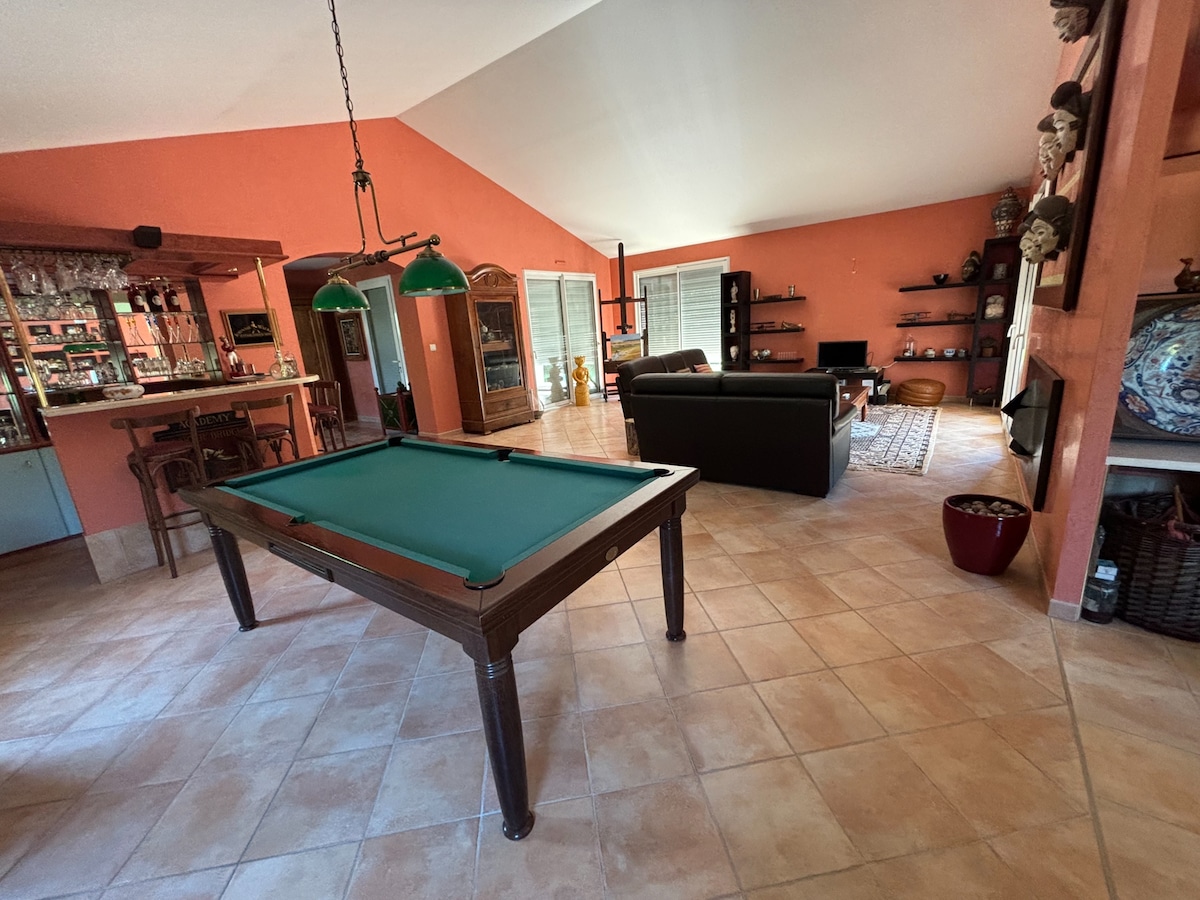An open living area is shown, featuring a green billiard table in the foreground. The warm orange walls complement the spacious layout, which includes a bar area, a comfortable sofa, and shelves with decor. Soft lighting enhances the inviting ambiance of the room.
