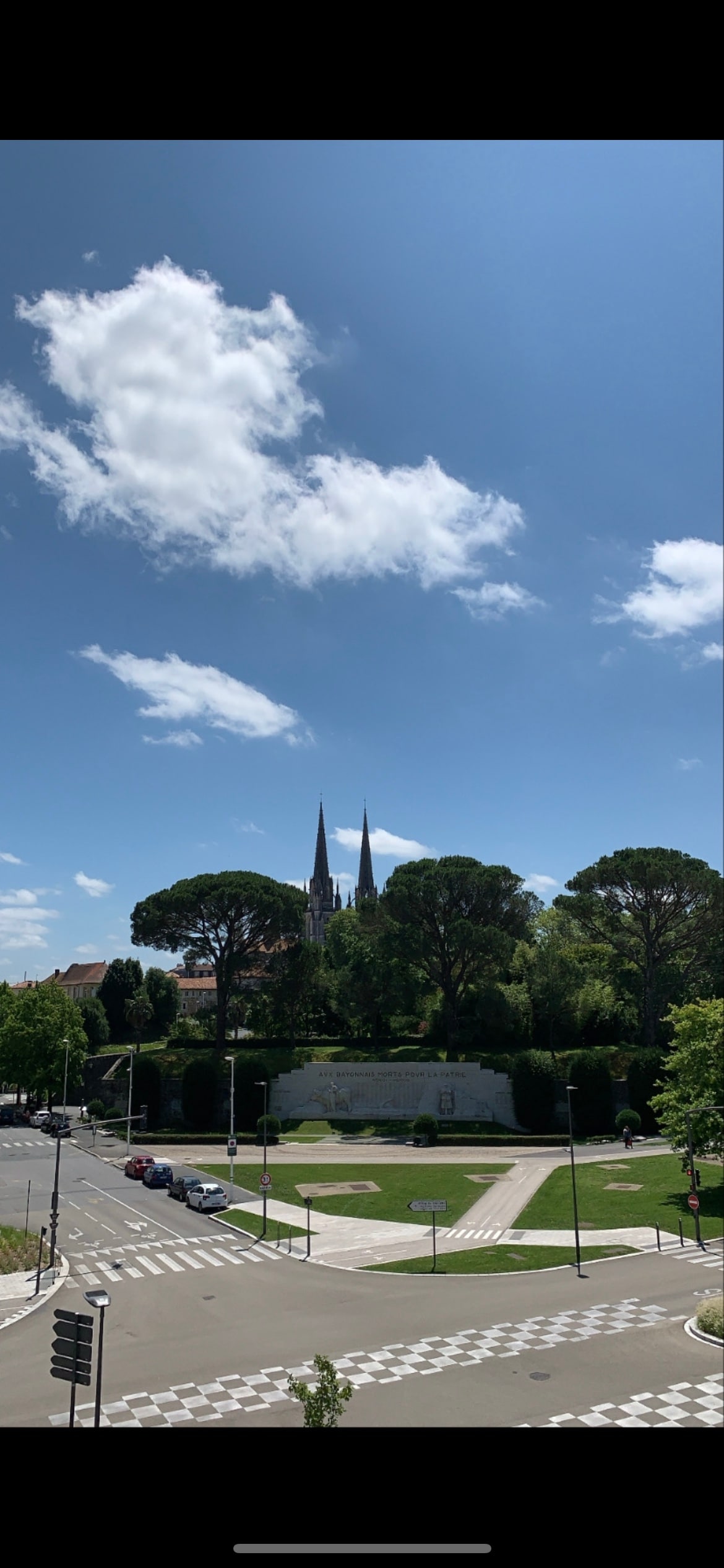 A clear blue sky is visible above the skyline, with two spires rising prominently among green trees. The view includes a well-maintained green space and a wide avenue leading towards the foreground, creating a sense of openness and tranquility.