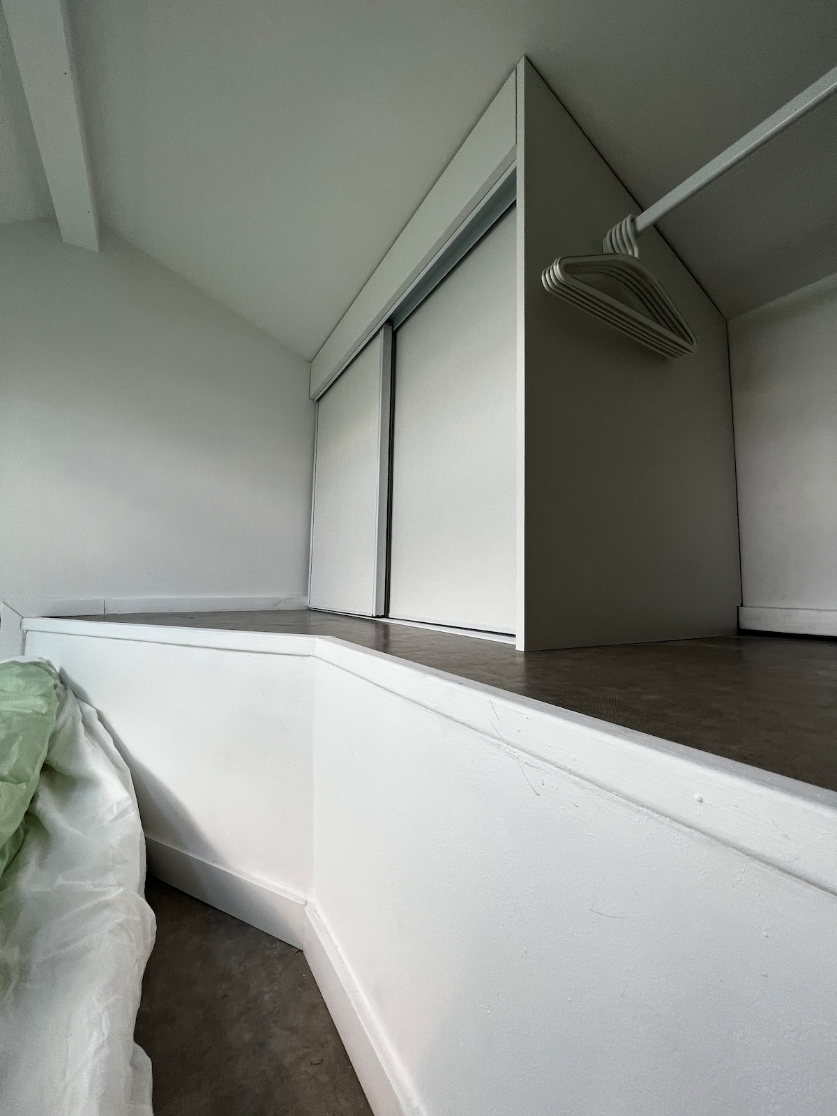 A corner area is presented with a built-in shelf and a smooth surface, featuring a soft green bedspread draped over the edge. Light enters through a partially closed window, creating a serene atmosphere in the space.