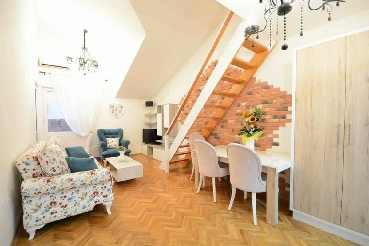 A bright living area features a floral-patterned sofa paired with a blue armchair. Light wooden flooring complements a white dining table surrounded by upholstered chairs. A rustic brick accent wall adds visual interest, while natural light pours in through a nearby window beneath a sloped ceiling.