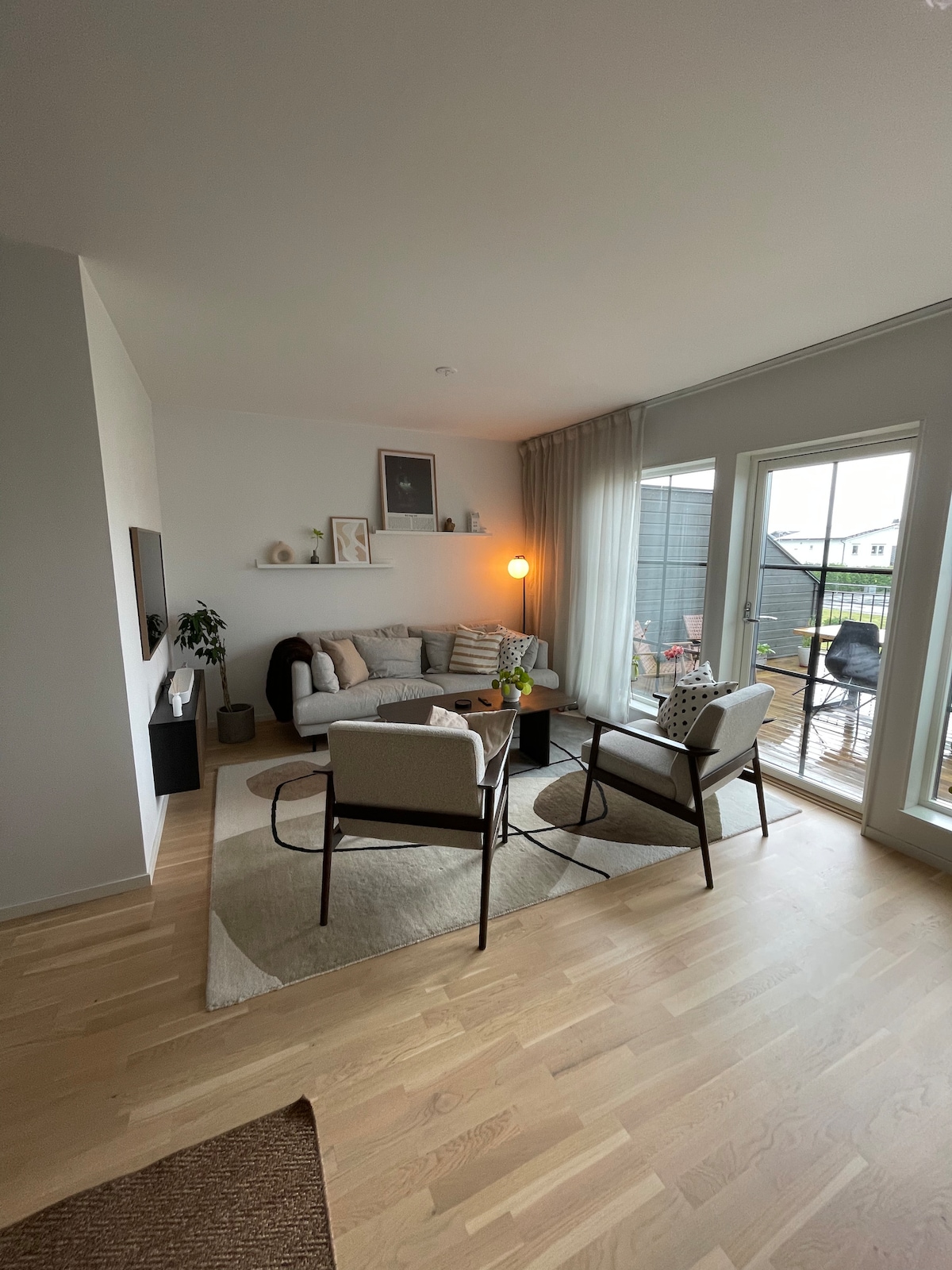 A bright living area is presented with modern furniture, featuring a light-colored sofa adorned with pillows. Two armchairs are positioned around a small table, while large glass doors provide access to a wooden terrace. Soft natural light enhances the space.