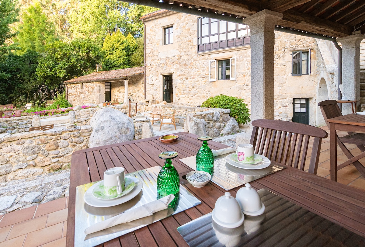 An outdoor dining area is set on a wooden table, featuring green glassware and white dishware. A stone facade of the house is visible in the background, along with a garden area filled with colorful flowers and well-maintained landscaping.