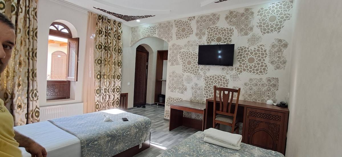 The room features two spacious beds with patterned covers, complemented by wooden furnishings. A flat-screen TV is mounted on the light-colored wall, enhancing the modern design. Elegant curtains frame the arched window, allowing natural light to fill the space.