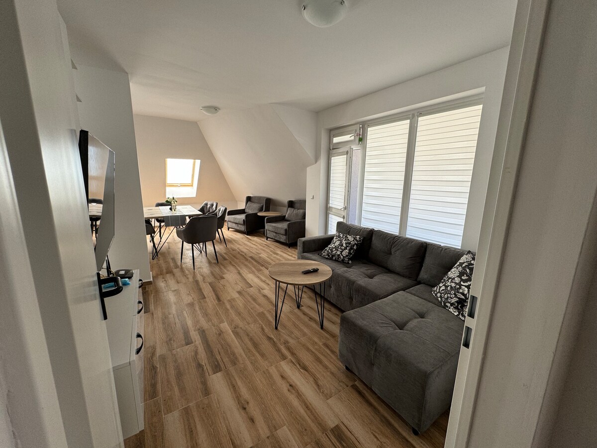 A spacious living area features large windows allowing natural light. Two comfortable sofas are arranged around a circular coffee table, while a dining table and chairs occupy the space adjacent. A TV unit is positioned on a wall opposite the seating area, enhancing the room's functionality.
