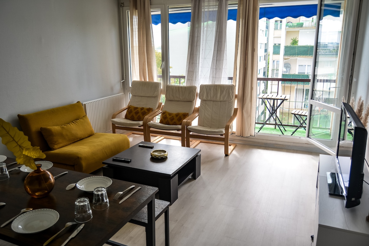 A bright living area features large windows dressed with light curtains, allowing natural light to fill the space. Three comfortable chairs are positioned next to a small outdoor balcony, complemented by a cozy sofa. A dining table is set with dinnerware, enhancing the inviting atmosphere.