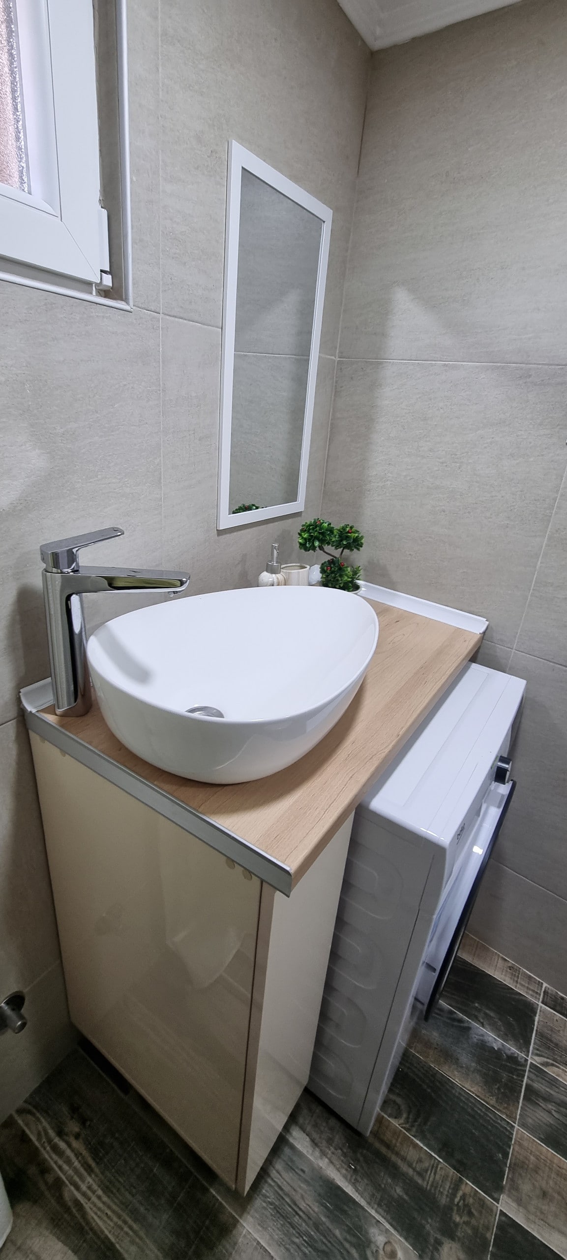 A modern bathroom features a sleek, white vessel sink positioned on a light wood countertop. A small mirror is mounted above the sink. The area includes a washing machine integrated beneath the countertop, and a small potted plant adds a touch of greenery.