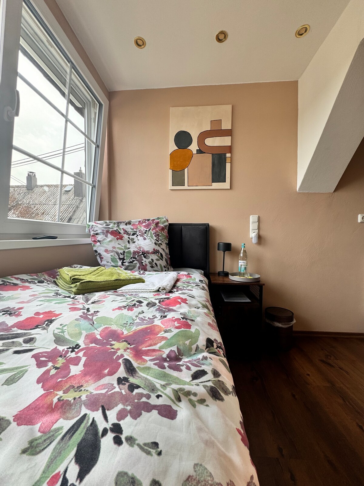 A cozy bedroom is presented with a spacious bed adorned in floral bedding. A small nightstand is positioned beside the bed, featuring a bottle of water. Natural light filters through a large window, casting a soft glow on the warm-toned walls and contemporary wall art.