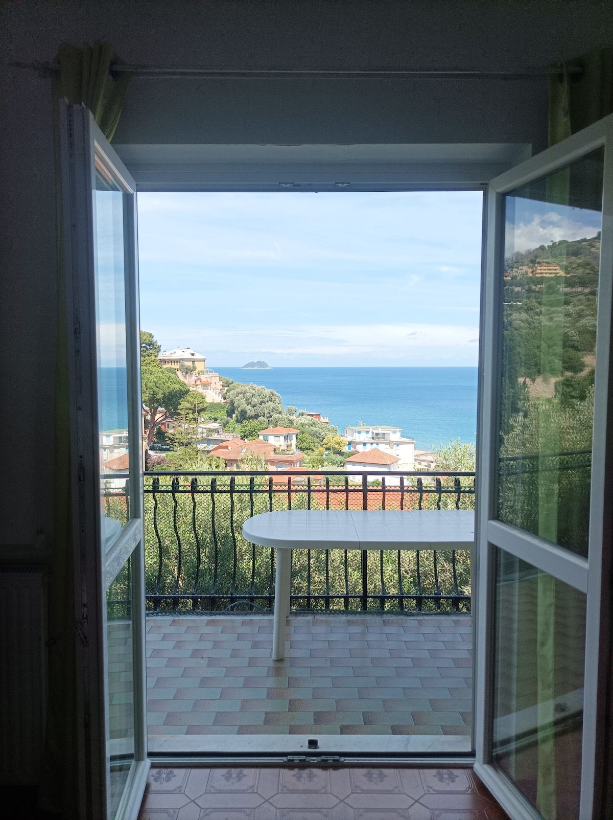 A view from an open set of double doors reveals a serene coastal landscape with the sparkling blue waters of the gulf and glimpses of colorful houses nestled among greenery. A small white table is positioned on the balcony, inviting relaxation in this tranquil setting.
