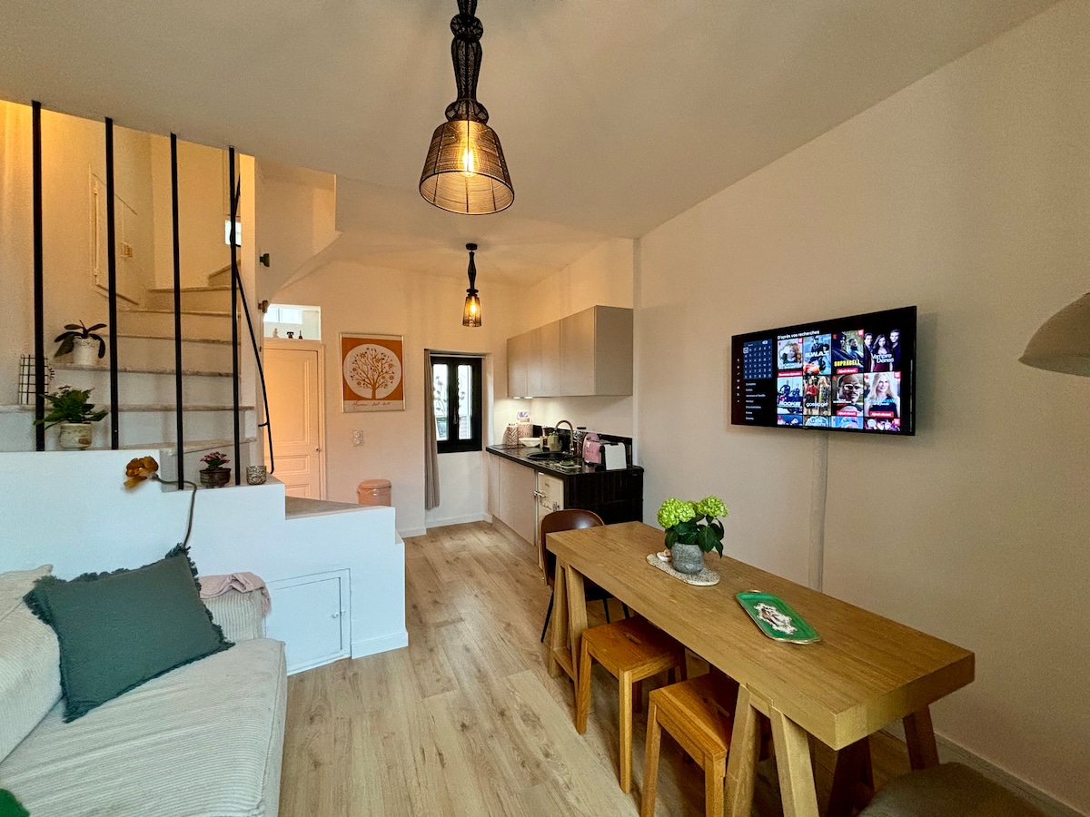 A spacious living area is highlighted by a wooden dining table accompanied by several chairs. Soft furnishings create a comfortable seating arrangement. A flat-screen TV is mounted on the wall, and a staircase leads to the upper levels, while light fixtures add a warm glow.