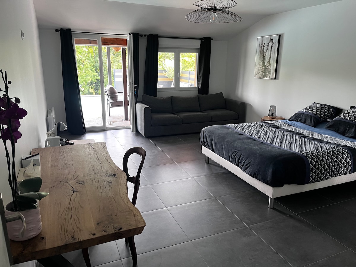 A spacious and light-filled room features a comfortable bed with patterned bedding and a cozy sofa. A wooden desk sits beside the window, and large glass doors open to a peaceful outdoor area, allowing natural light to enhance the modern decor.