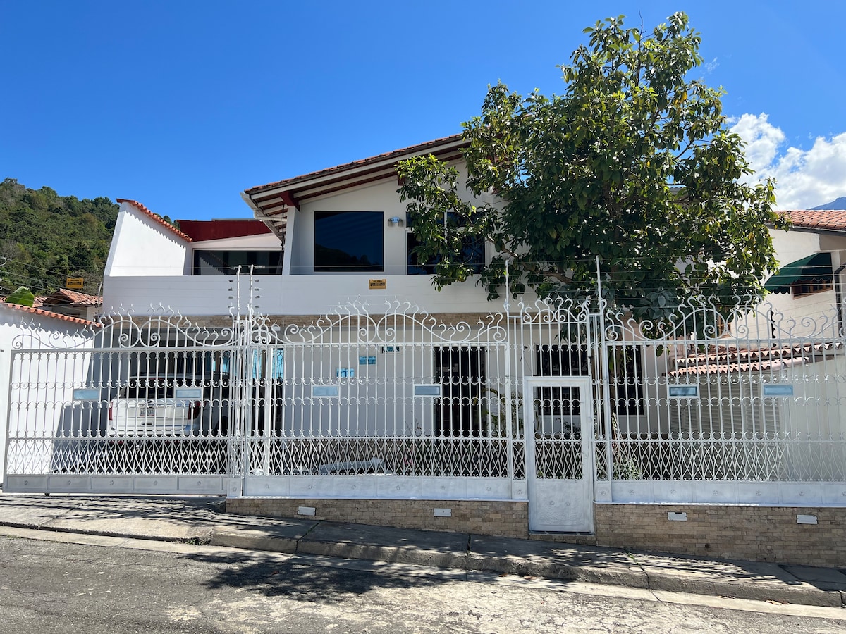 The exterior of a two-story house is visible, framed by a white decorative fence. A large tree provides shade near the entrance. The building features multiple windows and a welcoming facade under a clear blue sky, highlighting the tranquil neighborhood setting.