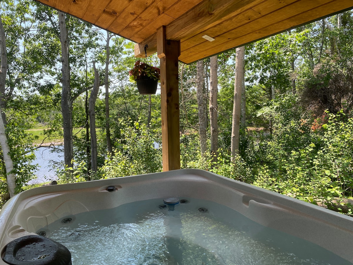 A hot tub is situated under a wooden structure, surrounded by lush green trees. The serene water reflects the natural scenery, with glimpses of the river visible beyond the foliage. A hanging planter adds a touch of nature to the space.