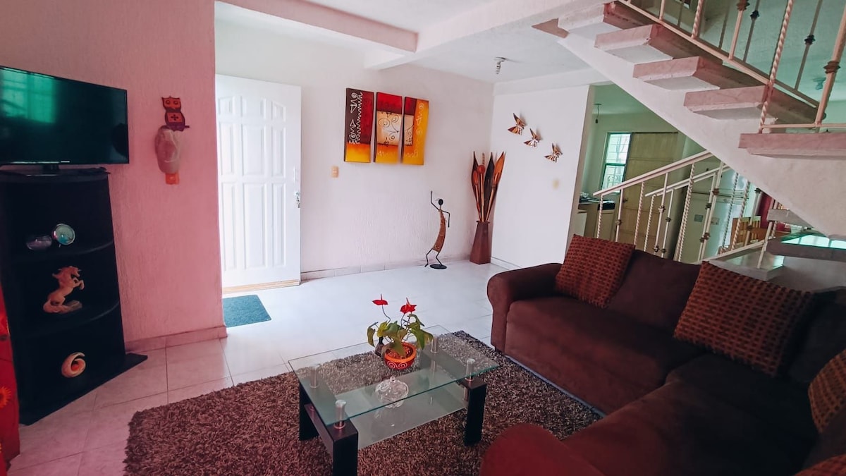 A spacious living area features a comfortable brown sofa facing a glass coffee table with a decorative plant. A wall-mounted television is positioned beside the entrance, and artwork with warm tones is displayed on the wall, contributing to an inviting atmosphere.
