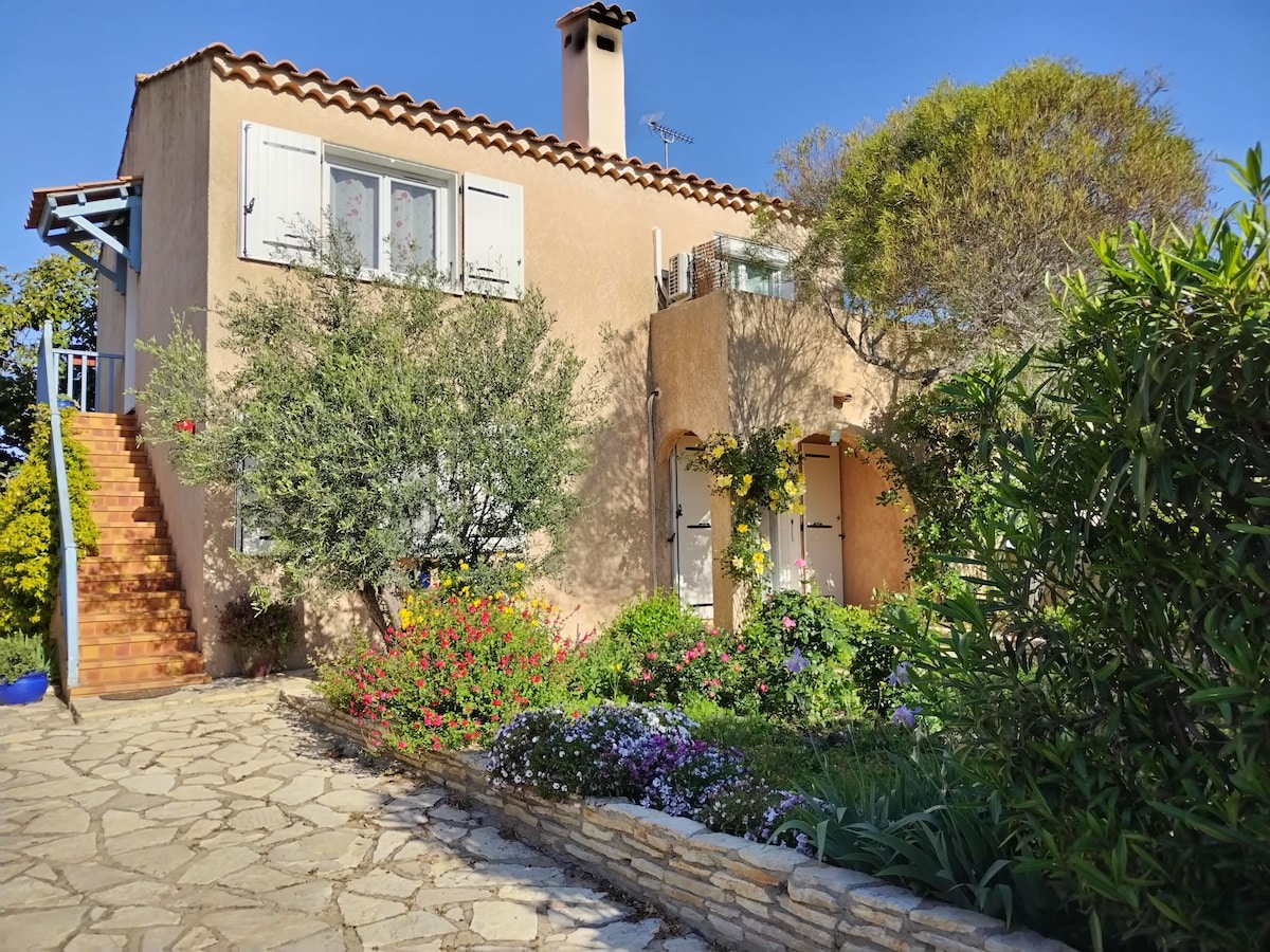 The exterior of a two-story house is surrounded by a lush garden featuring colorful flowers and greenery. Stone pathways lead to the entrance, and a staircase ascends to the upper level. Bright blue skies complement the peaceful atmosphere of the residential area.