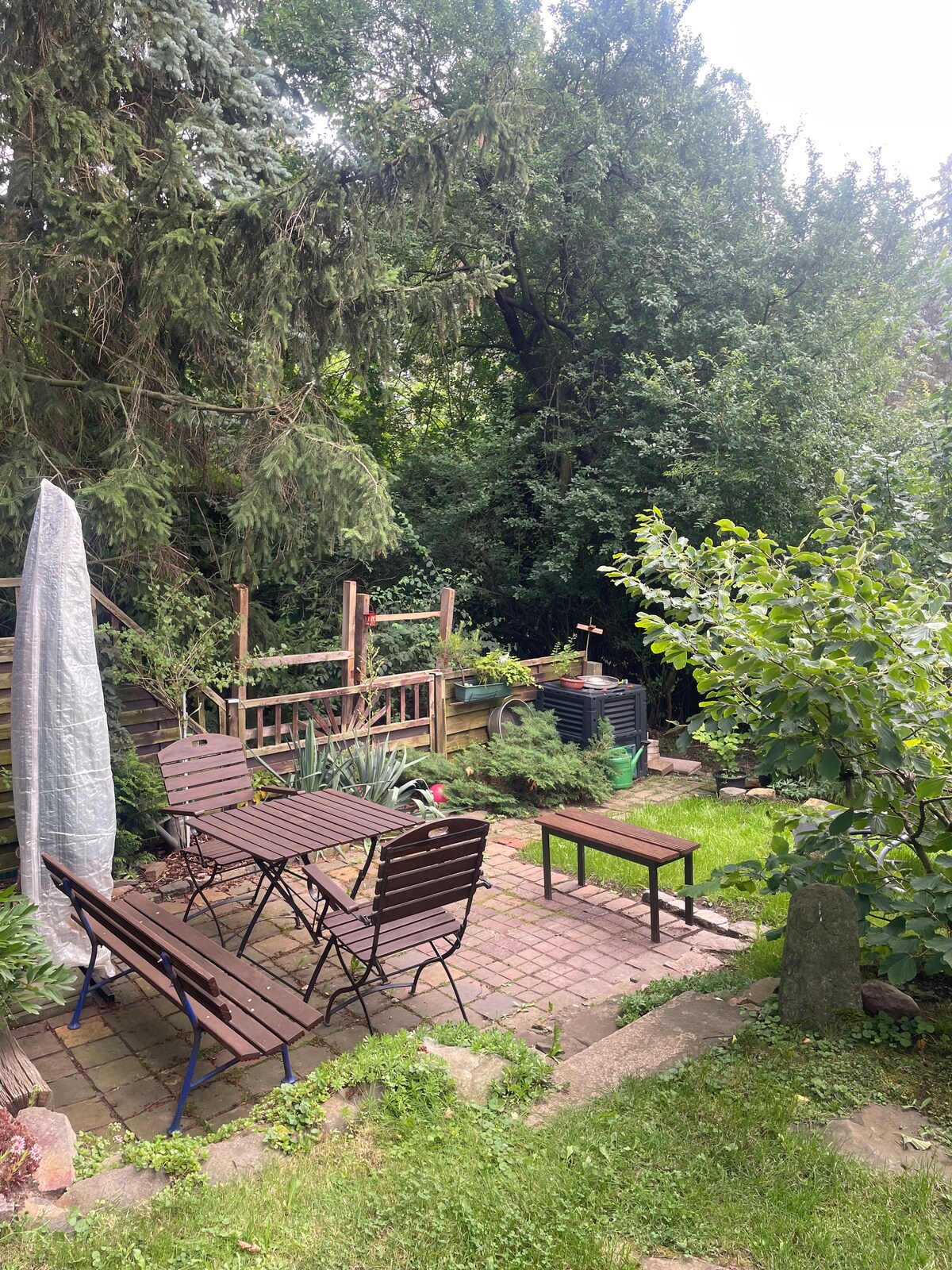 A secluded garden space features a brick patio framed by lush greenery. A set of brown outdoor chairs and tables is accompanied by vibrant plant life. A wooden deck leads up to a higher area, enhancing the feeling of intimacy and nature.