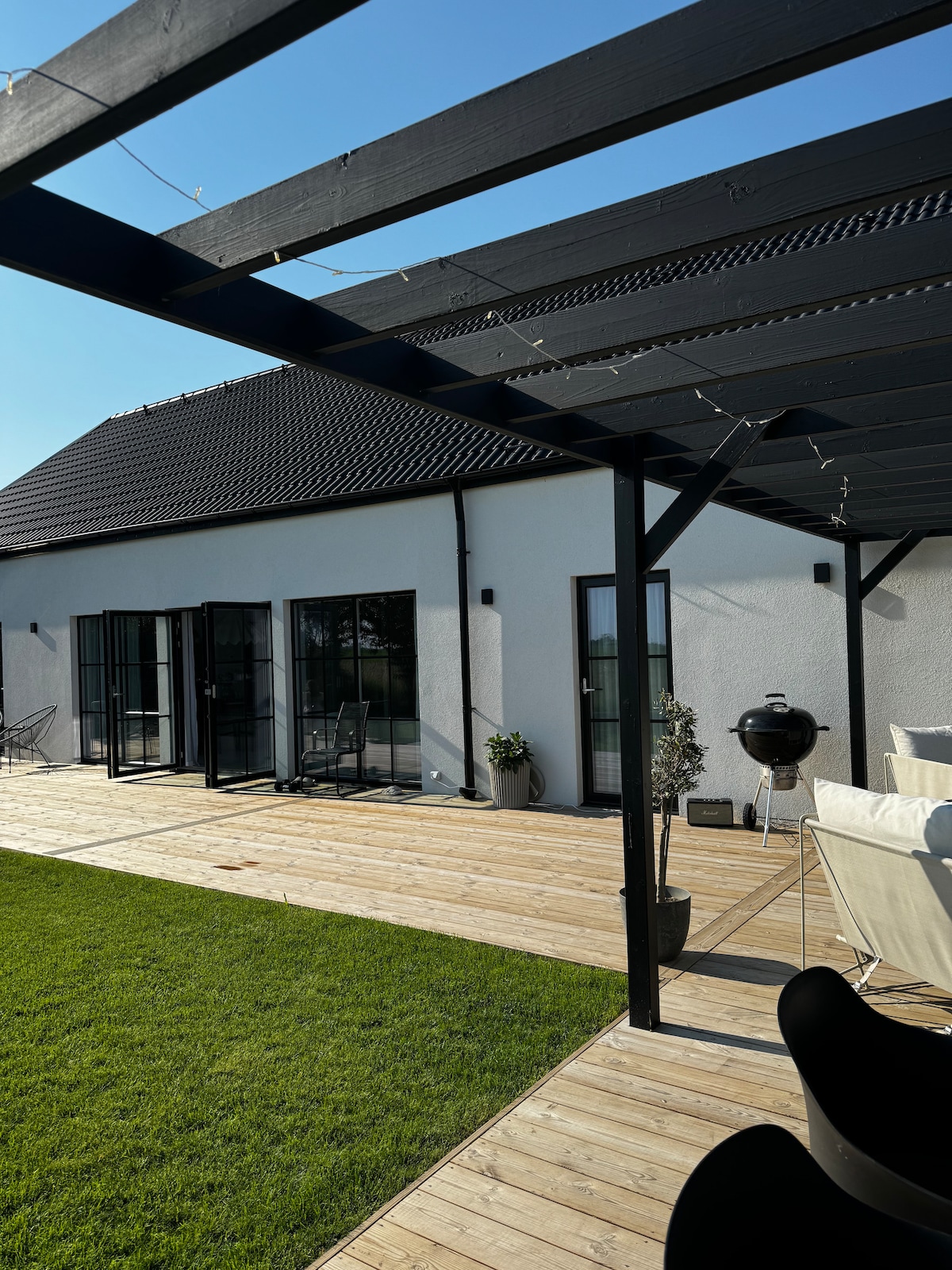 A spacious wooden deck is shown, framed by a black pergola. The area features green grass and includes a barbecue grill. Large glass doors lead into the villa, while a few outdoor seating options are arranged for relaxation.