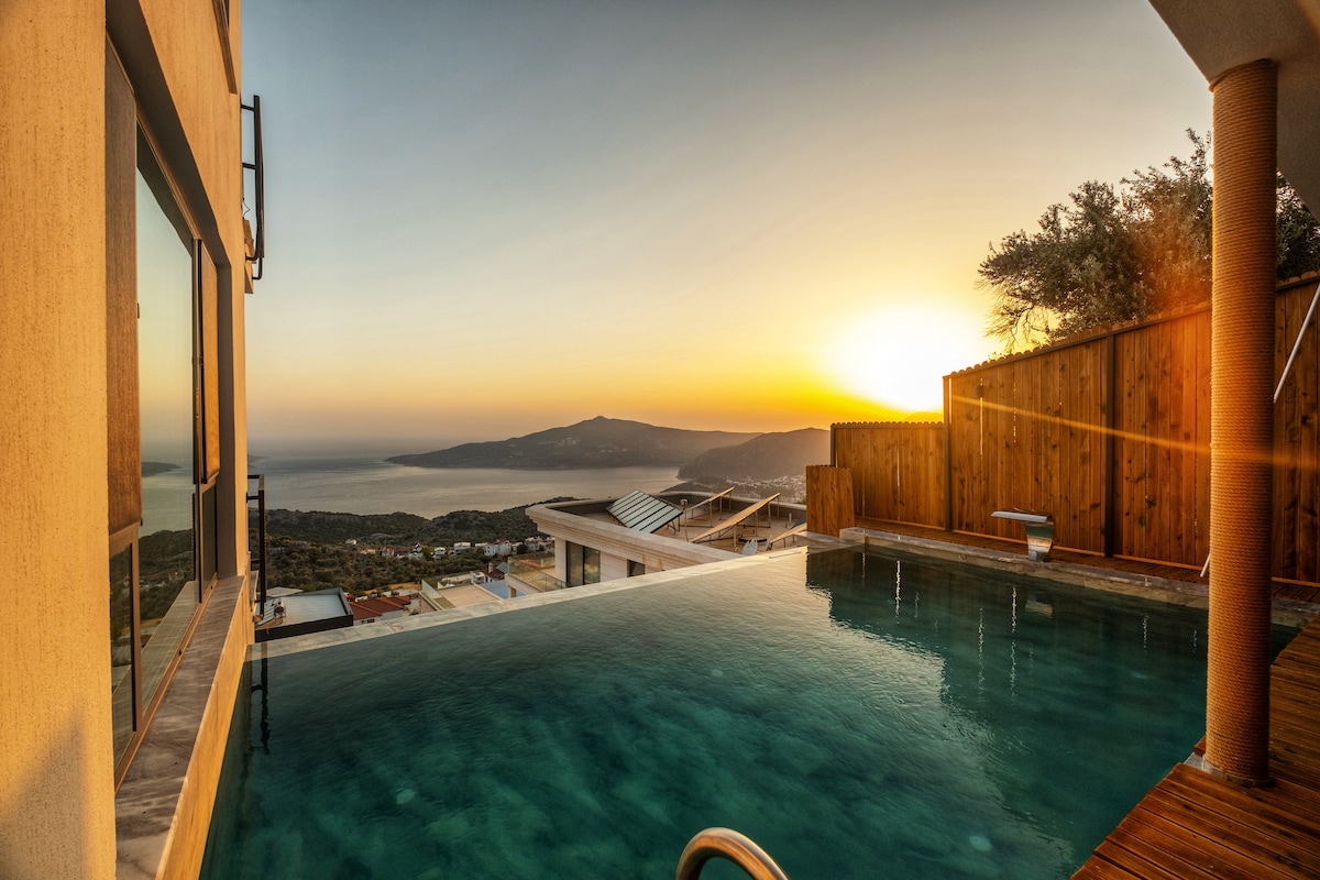 An infinity pool overlooks a stunning sunset over the sea, blending seamlessly with the horizon. The warm glow of the setting sun reflects off the tranquil waters of the pool, while surrounding trees and wooden fences offer a sense of privacy and nature.