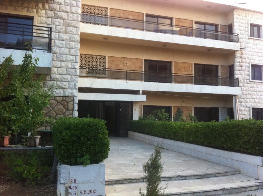 The exterior of a multi-level apartment building is displayed, featuring a stone facade and multiple balconies. A well-maintained pathway is lined with greenery, including shrubs and trees, leading towards the entrance. Large windows provide plenty of natural light.