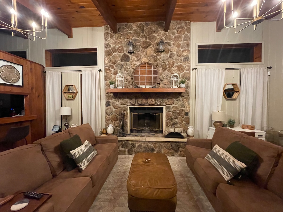 The living area features a rustic stone fireplace surrounded by soft seating and cozy decor. Dual windows allow natural light to brighten the space, while a coffee table sits at the center. Warm wood accents are highlighted by stylish light fixtures overhead.