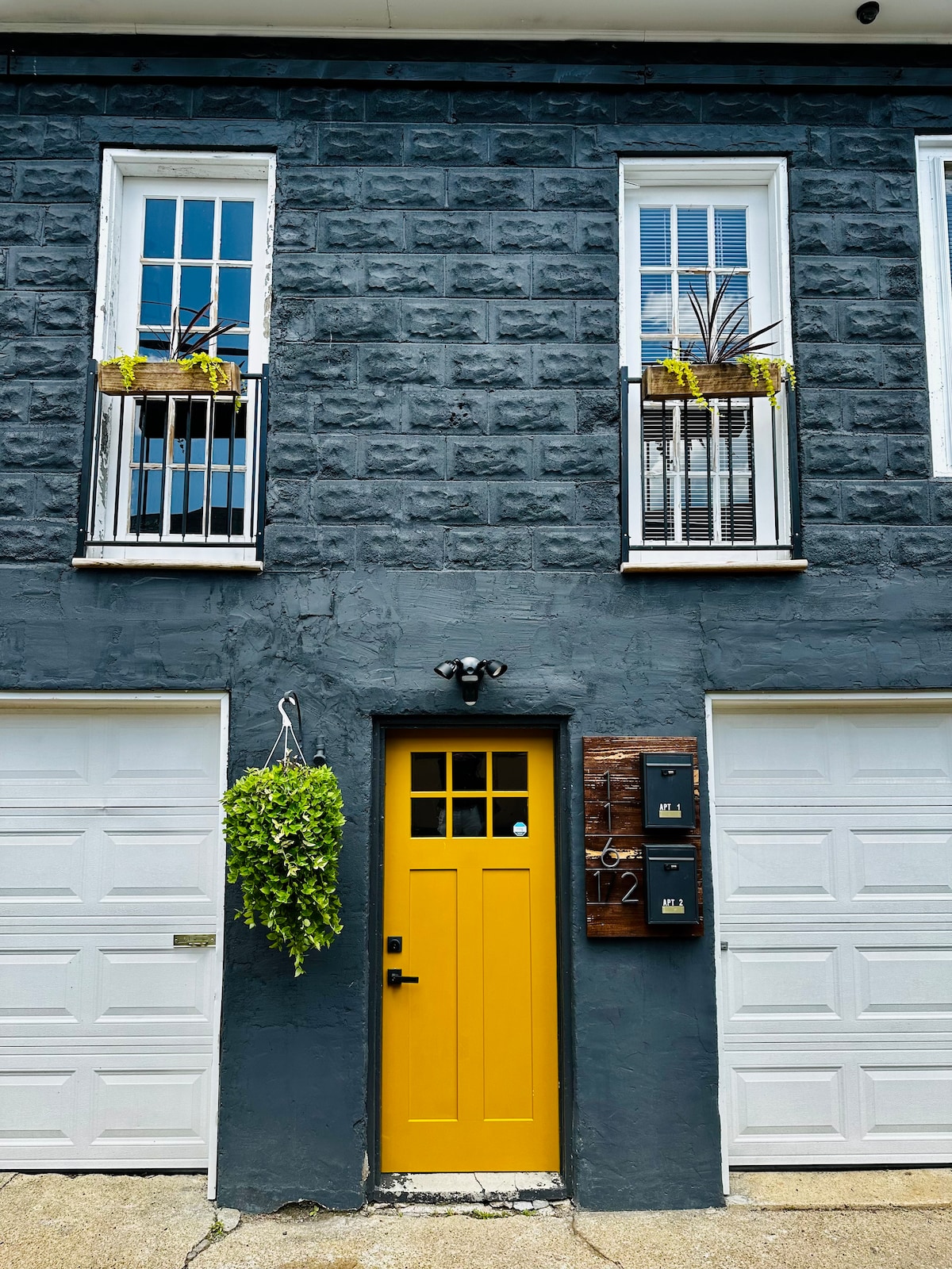 A vibrant yellow door stands at the center of a textured gray facade. Above the door, two windows are framed by black railings with planters. Informal greenery hangs beside the entrance, adding a welcoming touch to the exterior. Two garage doors are positioned below the windows.