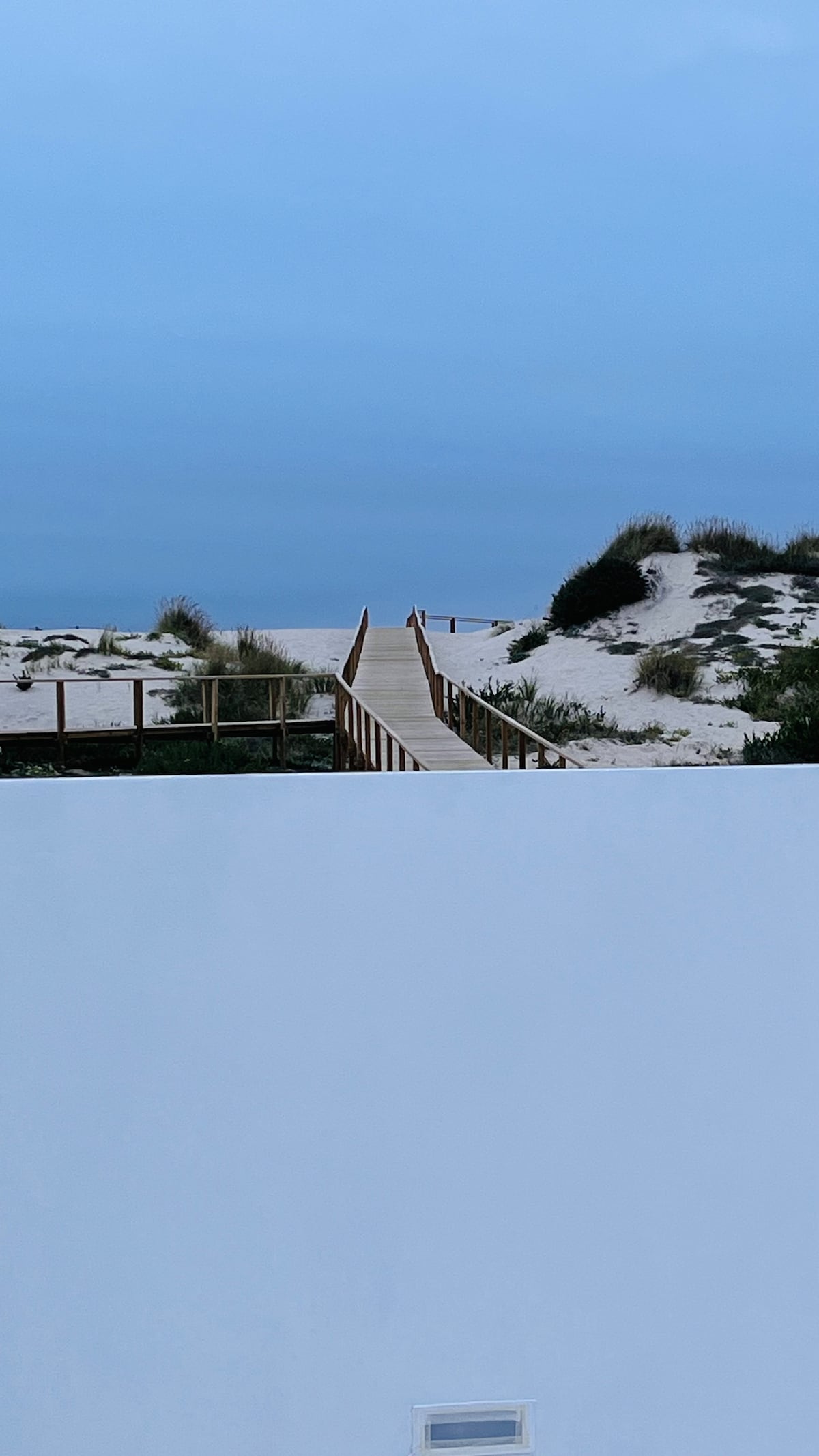 A wooden walkway extends through sandy dunes, leading toward the ocean. The pathway is bordered by patches of grass, and the soft colors of the sky suggest early evening or dawn. The scene conveys a serene coastal setting.