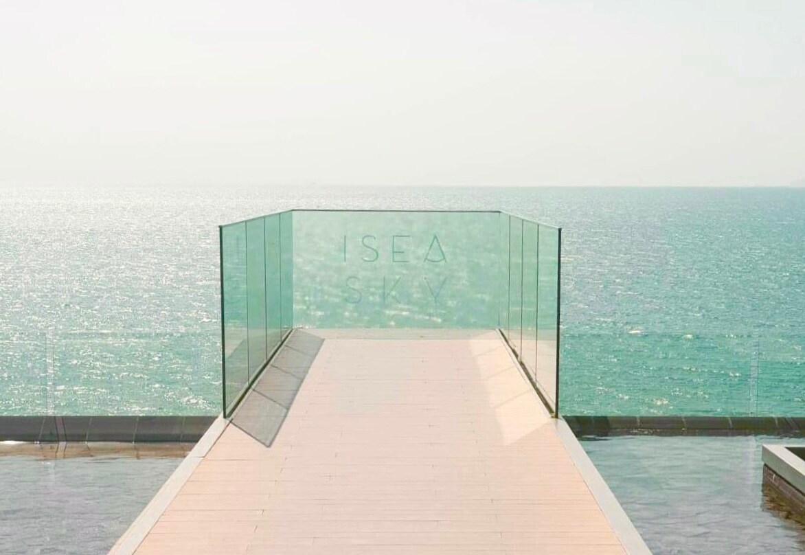 A sleek deck extends toward the ocean, bordered by transparent glass railings. The shimmering sea is visible in the background, reflecting soft light, while the clear sky creates a serene atmosphere. The deck offers an unobstructed view, inviting relaxation and contemplation.